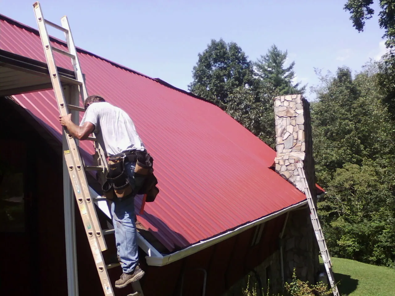 Roofer on a ladder beside a red metal roof and stone chimney — Soffit & Fascia Repair by Roofing Brothers in Mechanicville